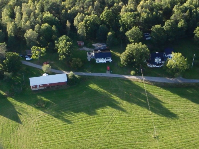 Flygbild över ett bostadsområde med ett hus nära en röd bil, gröna träd och fält.