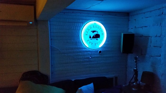 Rund ljusskylt med retrobil och text "Kees Kustom Est. 2003" belyst i blått på en vit trävägg inomhus.