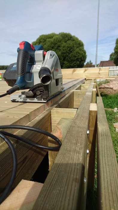 En elektrisk cirkelsåg placerad på nybyggd trädäckstrappa under arbete, med träd i bakgrunden.
