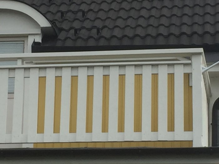 Vitt balkongräcke med vertikala gula och vita plankor under ett svart tak.