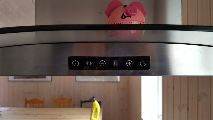 Kontrollpanel på köksfläkt med symboler för ström, belysning, och fläkthastighet under en röd timer-grisfigur.