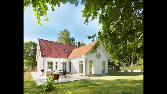 Vit villa med rött tak, veranda och trädgård, belägen i en solig, lummig miljö.