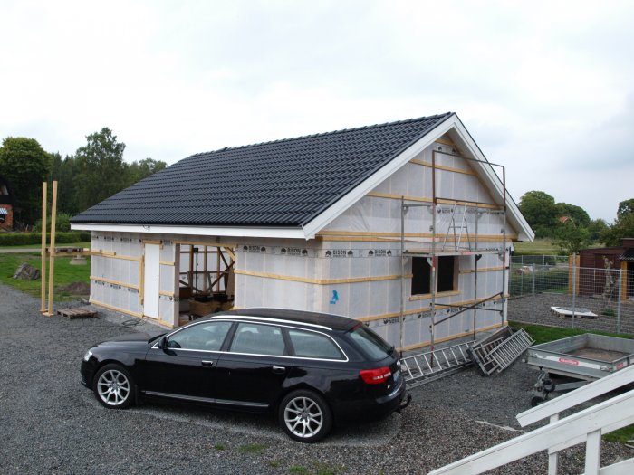 Hus under konstruktion med osynlig platta, omgiven av byggmaterial och en svart bil framför.