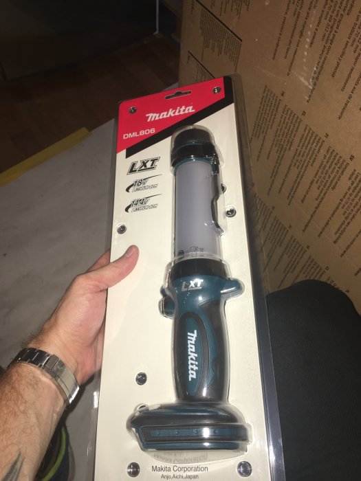 Hand som håller en ny Makita DML806 LED-lampa i originalförpackning, mot en bakgrund av kartonger.