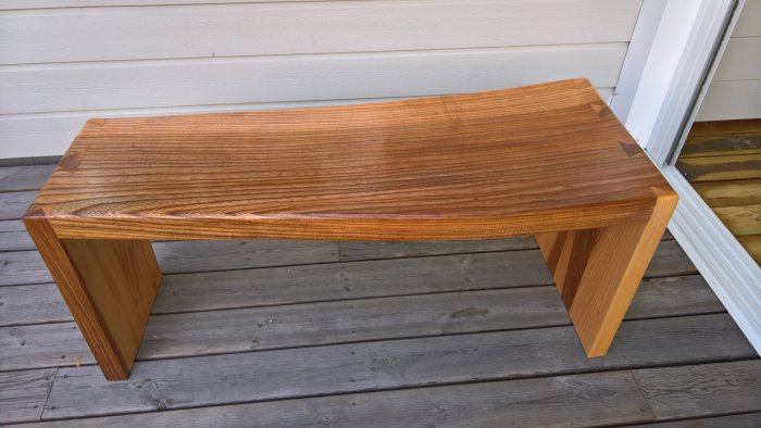 Handgjord bänk av böjd almplanka med finish av Danish Oil på en träaltan.