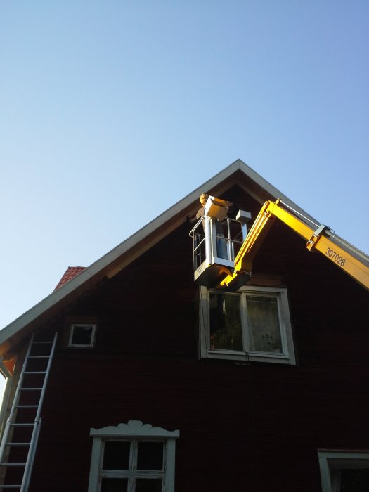 En person i en bomlift reparerar vindskivor på ett rött hus med stege nedanför.