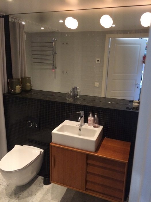 Renoverat badrum med svart kakel, spegel med belysning, träfärgat skåp och vitt duschdraperi.
