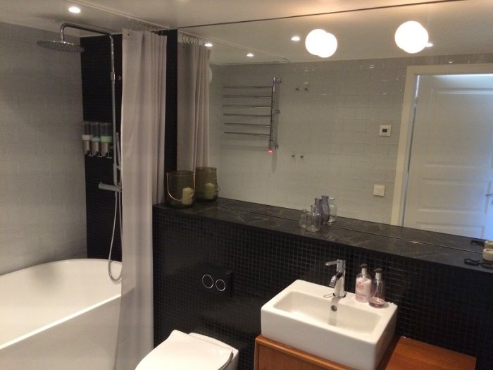 Renoverat badrum med svart kakel, måttbeställd spegel med belysning, dusch med draperi, tvättställ och badkar.
