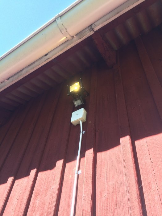 Ljus tänd på nyinstallerad utomhusarmatur med kablar längs en rödmålad vägg och takränna.