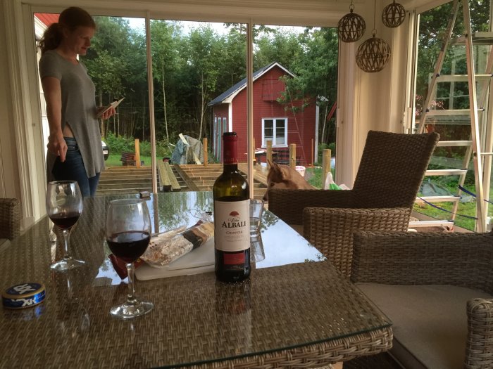 Utsikt från uterum med trägolv och pärlspont, kvinna ser på altanbygge, vin på bordet.