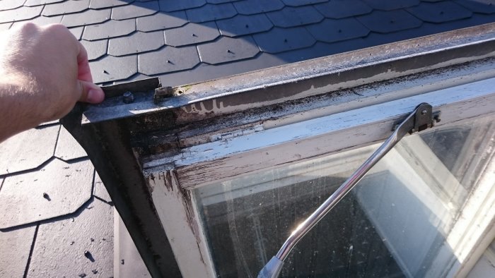Närbild på handen som håller i kanten av ett slitet takfönster med flagande färg och synliga gångjärn.