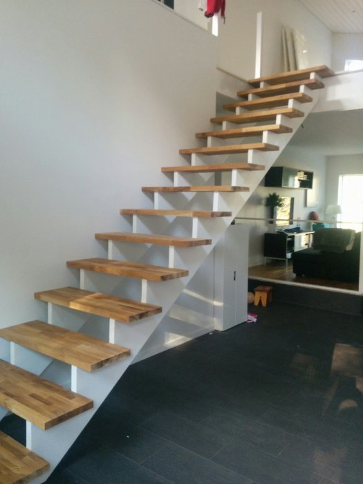 Svart flislagt gulv med en moderne, flytende trapp i tre og metall, leder opp til en lys loftsetasje i en boliginnredning.