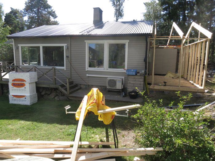 Tillbyggnad av fritidshus med trästomme och byggmaterial i förgrunden, befintligt hus med nock i bakgrunden.