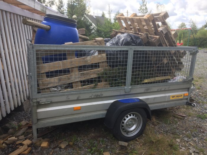Släpvagn fullastad med bråte som träplank och blå tunna, parkerad med hjulet i en grop vid en trävägg.