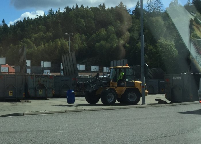 Gul hjullastare framför återvinningsstationens container med metallavfall, blå tunna och skog i bakgrunden.