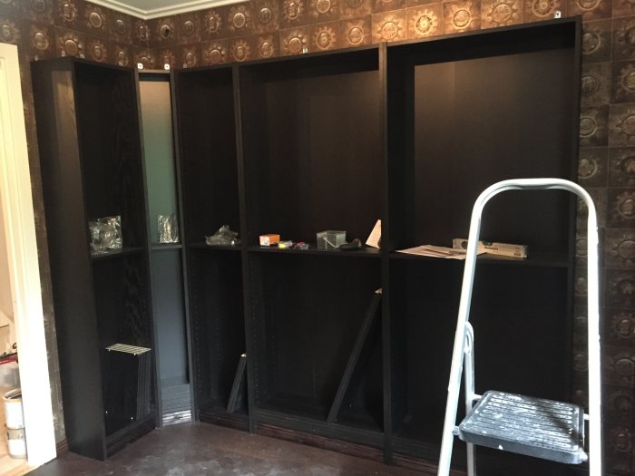 Svart bokhylla med en frostat glasdörr och en ofrostad, stege framför, i ett rum under renovering.