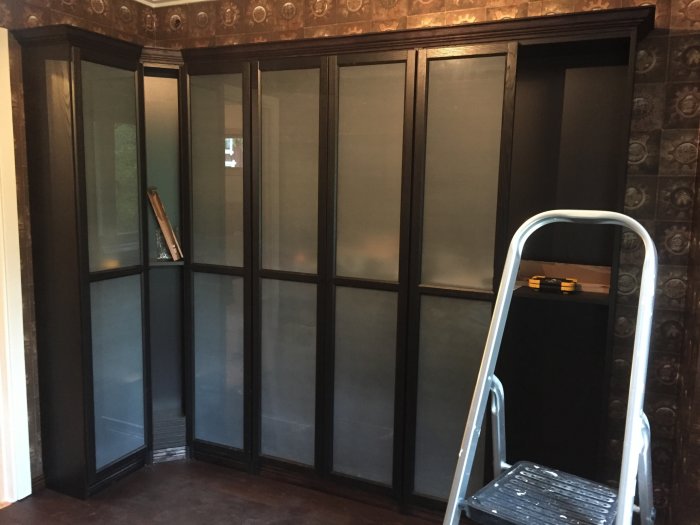 Bokhylla med frostat glas och en ofrostad dörr, stege och verktyg i ett rum under renovering.