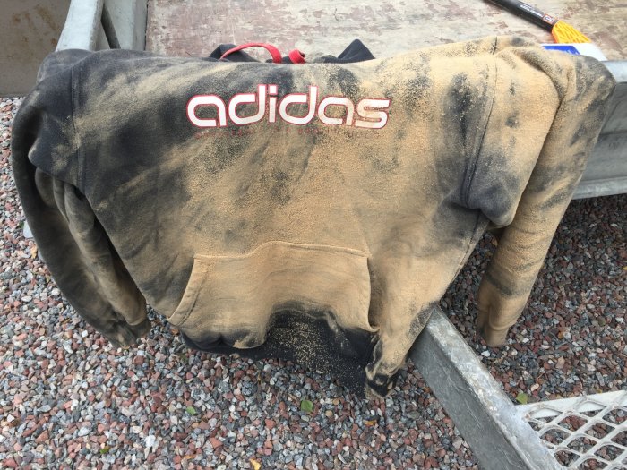 En svart Adidas-tröja täckt med mängder av mdf-sågspån, vilande på en grusig mark.