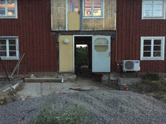 En fasad av ett hus under renovering vid skymning med öppen dörr, grushög och byggmaterial synliga.