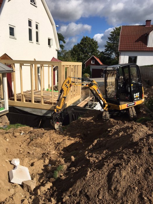 Grävmaskin återfyller jord runt källaren bredvid ett hus med pågående bygge av trädäck.