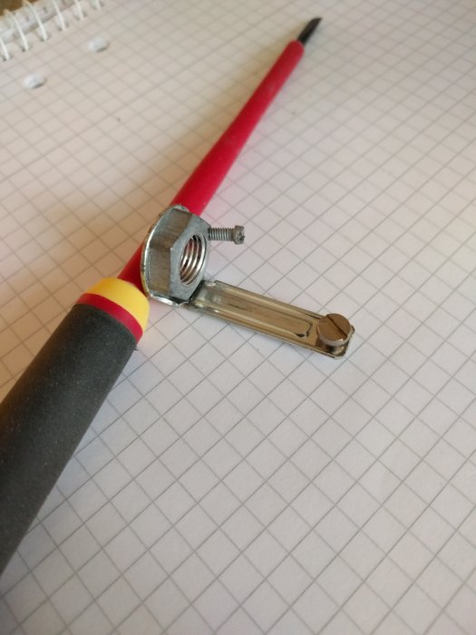 Metallklämma fästad på röd kabel med kulled och skruv på rutigt papper.