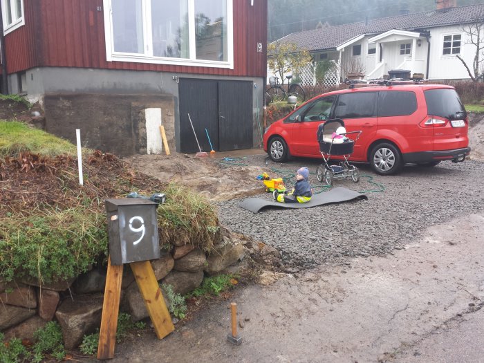 Grävt område framför hus med grus, verktyg och barnleksaker, en röd bil och en barnvagn syns.