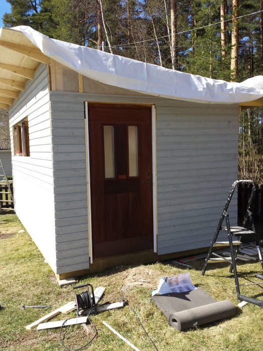 Nybyggd oisolerad friggebod med träfasad och tak under uppbyggnad, verktyg synliga framför byggnaden.