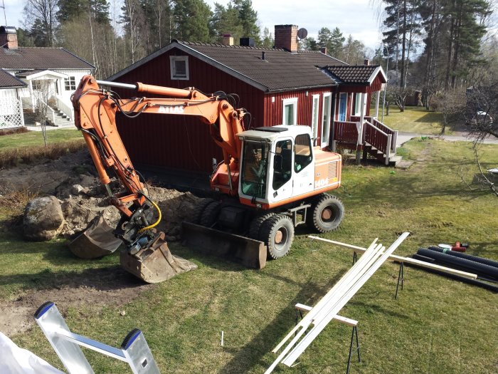 Grävmaskin framför röd villa under arbete med dränering och borttagen farstukvist, stora stenar synliga.