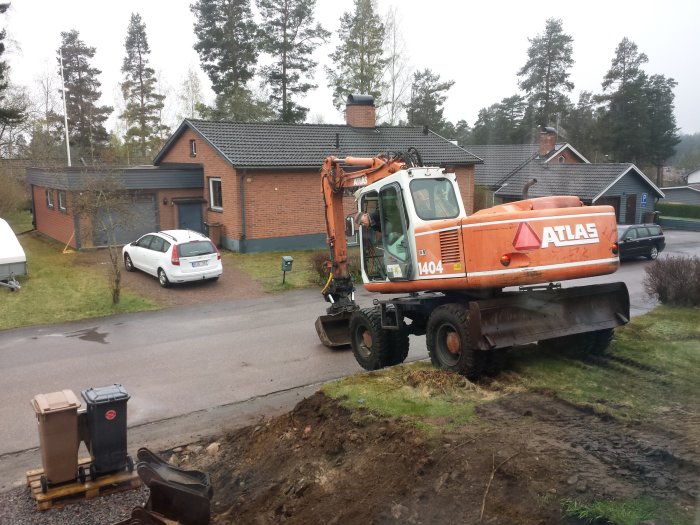 Grävmaskin framför hus under dräneringsarbete med synliga jordhögar och stenar.