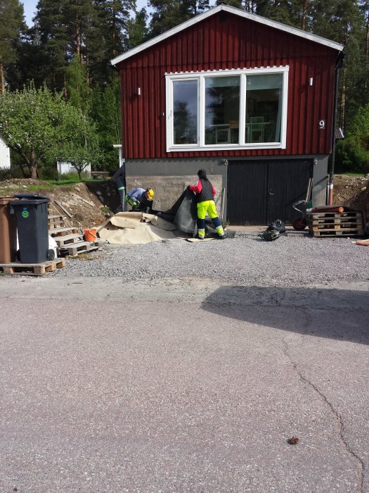 Arbetare utför dräneringsarbete vid husgrund med avlägsnad farstukvist och synliga stora stenar.