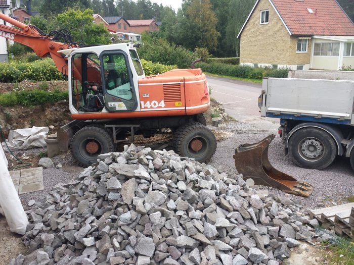 Grävmaskin bredvid en hög av stenar för gabioner i ett bostadsområde, med hus i bakgrunden.