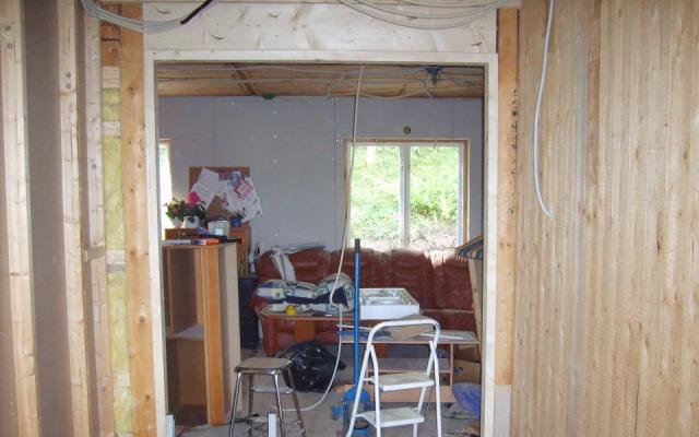 Rum under renovering med et åbent dørhul, stige og værktøj spredt omkring, samt en brun sofa og et vindue med udsigt til have.