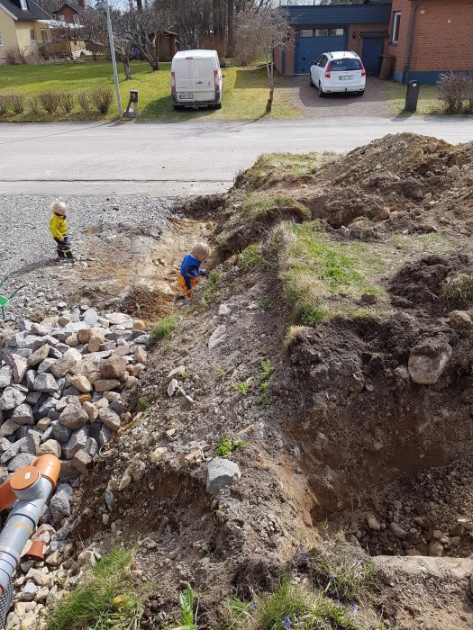 Två barn leker vid en stor grushög bredvid en grävd grop vid en bostadsdriveway.