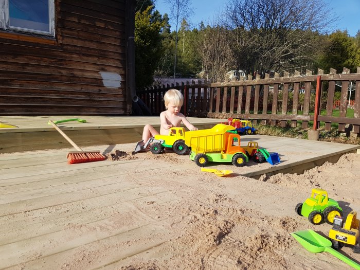 Barn leker i nybyggd sandlåda med leksakslastbilar och spadar på solig gård.