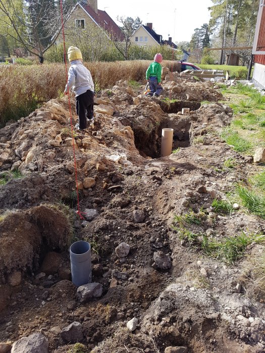 Grop för gjutning med sten och grus, rör i marken och två barn i bakgrunden.