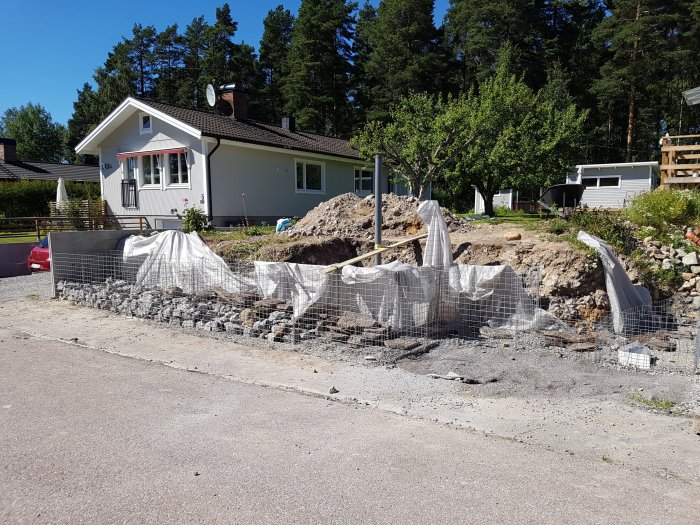 Gabionsten och nätburar på en husuppfart, arbete med grävning och anläggning av stödmur pågår.