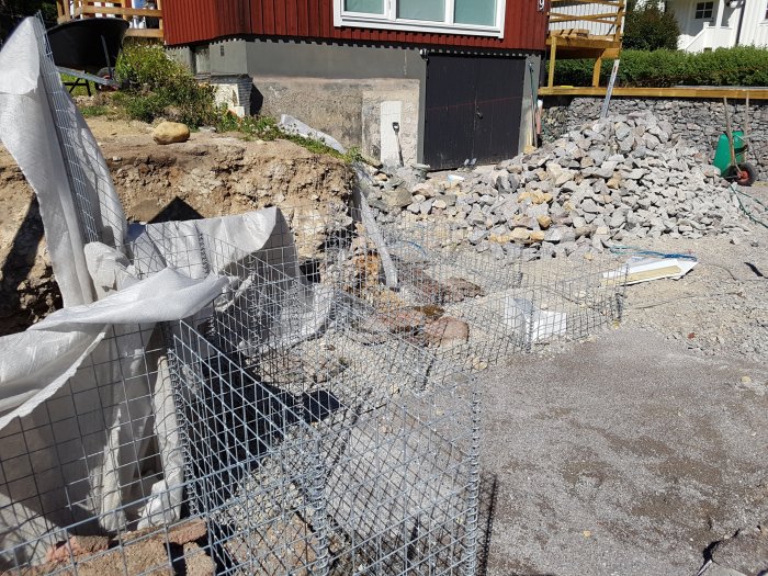 Byggplats med grushög, gabionnätburar och byggmaterial framför en röd villa.