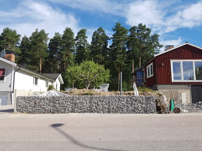 En gabionmur under konstruktion framför hus, halvmeter kvar till grannens L-stöd.