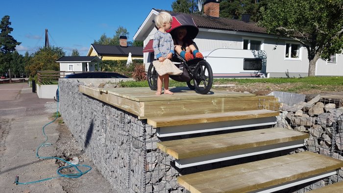 Två barn på en nyligen installerad trappa av trä med gatstenar undertill och hus i bakgrunden.