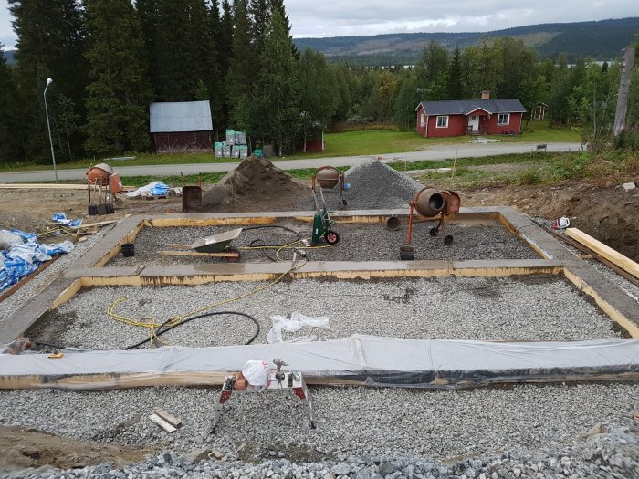 Grundarbete för byggprojekt med grus och formar, betongblandare och verktyg i förgrunden.