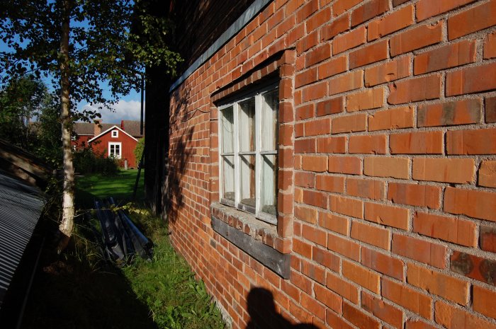 Delvis skuggad lada med röd tegelvägg och vitt fönster, skadat trä under fönstret, mot bakgrund av en röd stuga och grönska.