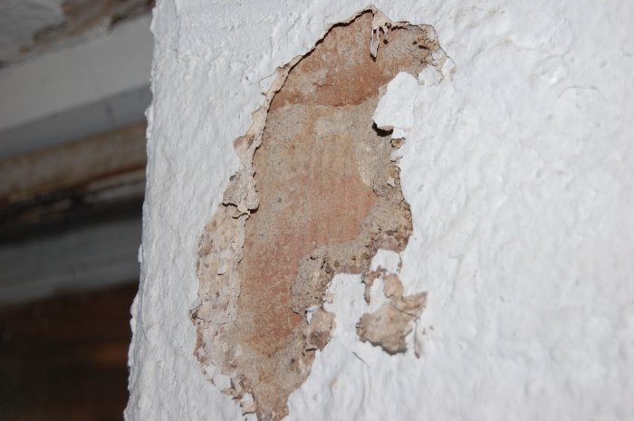 Närstående bild av en skadad verkstadsvägg med flagande ytbehandling och exponerat murverk vilket tyder på fuktproblem.