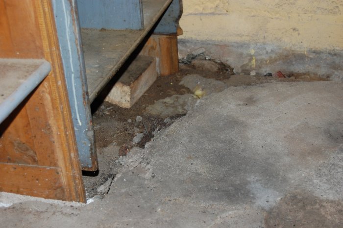 Skadat betonggolv i hörnet av en verkstad med synliga hål och sprickor nära en trätrappa och gul vägg.