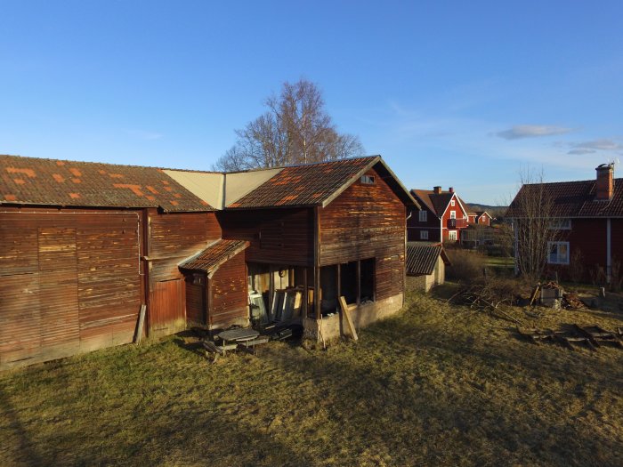 Gammal lada med öppen underdel och övervåning, omgiven av gräs och närliggande hus i dagsljus.