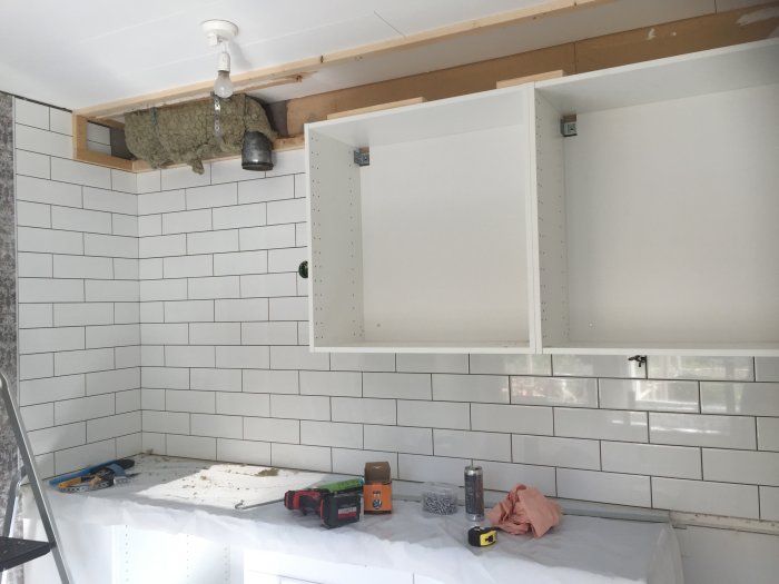 Kök under renovering med nybyggd regelram för täckskiva, vita kakelväggar och köksskåp utan dörrar.