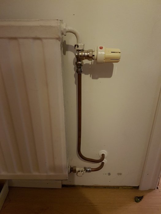 Vit radiator ansluten till kopparledningar med termostat och anslutningskopplingar monterade på vägg.