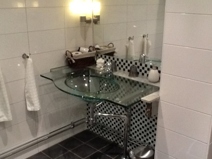 Genomskinligt glashandfat på metallställ med synligt vattenlås och svartvit kakelbakgrund i ett hotellbadrum.