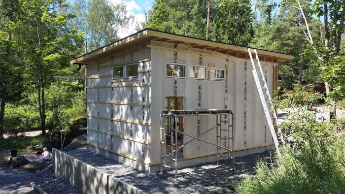 En byggplats för hus med isolermaterial synligt, omonterade plankor och ställningar, omgiven av grönska.