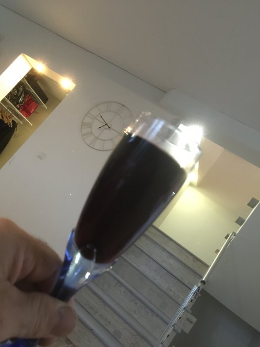 Hand håller ett champagneglas med röd dryck, otydlig bakgrund med en väggklocka och en stege.