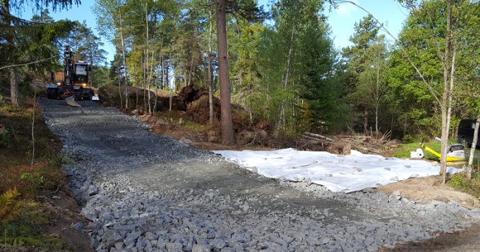 Grusväg upp på ett berg med nyanlagd parkeringsplats omgiven av skog och en grävmaskin i arbete.